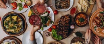 Plusieurs plats à partager sont disposés sur une tbale. DEs mains traversent cette table en se servant ou en trinquant avec des verres.