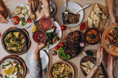 Plusieurs plats à partager sont disposés sur une tbale. DEs mains traversent cette table en se servant ou en trinquant avec des verres. - Agrandir l'image, fenêtre modale