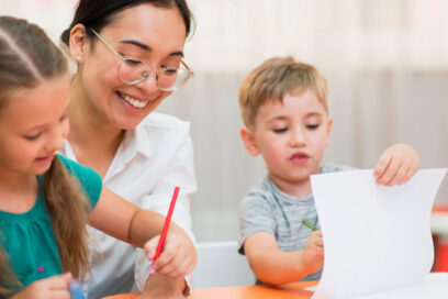 Une ATSEM, souriante et attentive, accompagne deux jeunes enfants assis à une table. Les enfants dessinent et manipulent des feuilles avec des crayons de couleur, dans un moment calme d’activité en classe. - Agrandir l'image, fenêtre modale