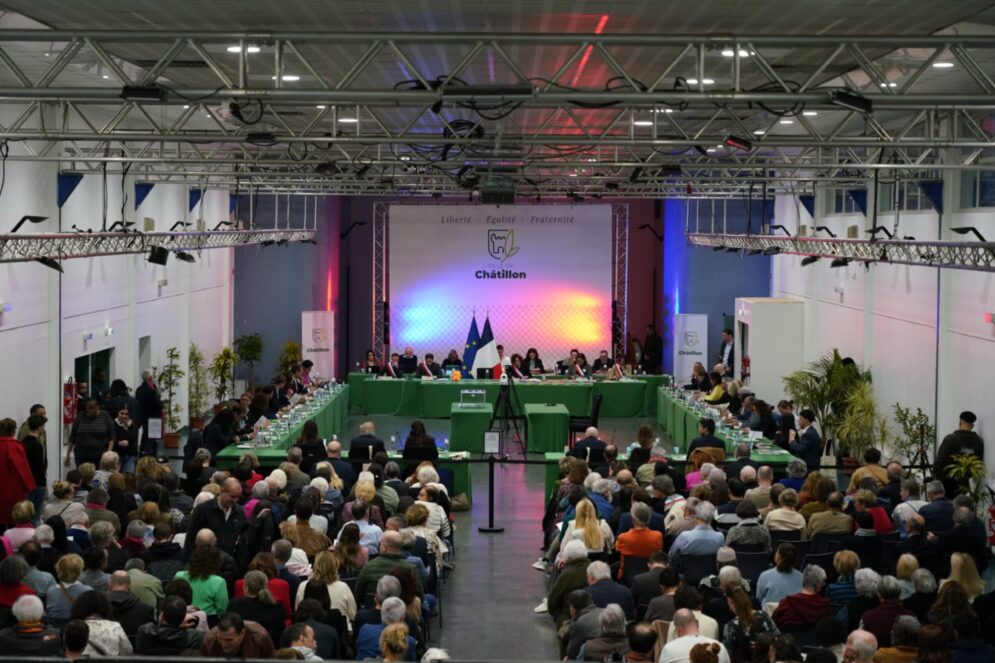 Vue large de la salle de l’Espace Maison Blanche lors de la séance d’installation du Conseil municipal de Châtillon : les élus sont installés en U autour de tables vertes, face au public nombreux assis dans la salle, avec les drapeaux français et européen au centre et le logo de la Ville en arrière-plan.
