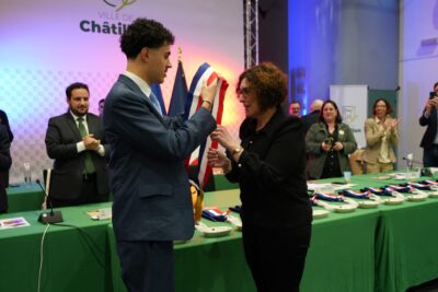 Jean-Baptiste Bénôt, jeune élu en costume, remet l’écharpe tricolore à Nadège Azzaz lors de la séance d’installation du Conseil municipal, sous les applaudissements des autres élus en arrière-plan. - Agrandir l'image 7 sur 9, fenêtre modale