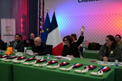 Autour de la table du Conseil municipal, Nadège Azzaz lève la main pour voter, entourée d’autres élus assis, avec des écharpes tricolores disposées devant eux et les drapeaux français et européen en arrière-plan. - Agrandir l'image 8 sur 9, fenêtre modale