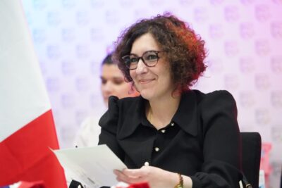Nadège Azzaz, assise à la table du Conseil municipal, lit un document avec un sourire, lors de la séance d’installation, avec le drapeau français visible au premier plan. - Agrandir l'image 9 sur 9, fenêtre modale