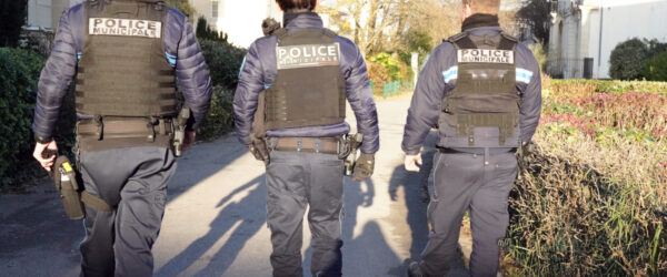 Trois agents de la Police municipale marchent côte à côte dans une rue résidentielle, vus de dos. En uniforme et équipés, ils effectuent une patrouille à pied entre des habitations et des espaces végétalisés.