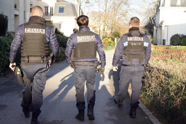 Trois agents de la Police municipale marchent côte à côte dans une rue résidentielle, vus de dos. En uniforme et équipés, ils effectuent une patrouille à pied entre des habitations et des espaces végétalisés.