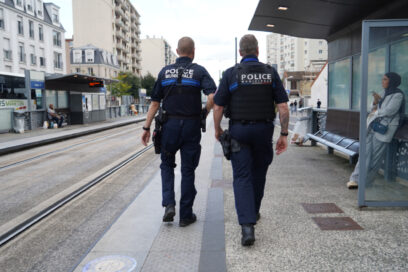 Deux agents de la Police municipale marchent côte à côte sur un quai de tramway en ville. Ils sont vus de dos, en uniforme, avec leur équipement. Autour d’eux, on distingue des immeubles, un abri de station et des passants. - Agrandir l'image, fenêtre modale