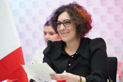 Madame la Maire, assise derrière une table, sourit en lisant un document qu’elle tient en main. Elle porte des lunettes et une veste noire. Un drapeau français est visible à ses côtés, dans un cadre officiel, avec une personne en arrière-plan. - Agrandir l'image, fenêtre modale