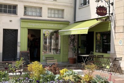 Façade vert tendre du salon de thé Bergamote & Grain de café, avec une devanture ouverte sur la rue, une petite terrasse équipée de tables et chaises pliantes, et des plantes fleuries au premier plan. Une enseigne indique “Salon de thé” au-dessus de l’auvent. - Agrandir l'image, fenêtre modale