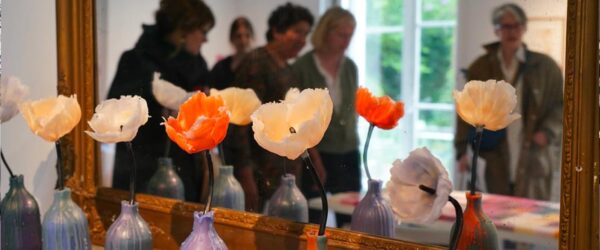 Vases colorés alignés sur une table en marbre, chacun contenant une fleur blanche ou orange, devant un grand miroir doré où se reflètent plusieurs personnes floues dans une pièce lumineuse.