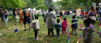 Foule de familles rassemblées dans un parc lors d’une fête en plein air : des enfants jouent avec des rubans colorés et des cerceaux pendant que des adultes les accompagnent, au milieu de stands, de guirlandes et d’animations installées sur la pelouse entourée d’arbres.