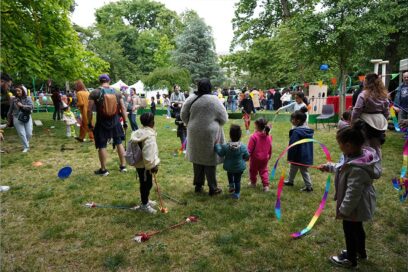 Foule de familles rassemblées dans un parc lors d’une fête en plein air : des enfants jouent avec des rubans colorés et des cerceaux pendant que des adultes les accompagnent, au milieu de stands, de guirlandes et d’animations installées sur la pelouse entourée d’arbres. - Agrandir l'image, fenêtre modale