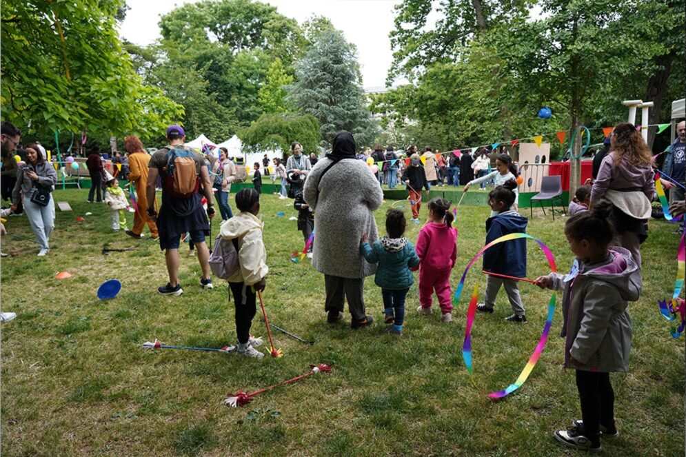 Foule de familles rassemblées dans un parc lors d’une fête en plein air : des enfants jouent avec des rubans colorés et des cerceaux pendant que des adultes les accompagnent, au milieu de stands, de guirlandes et d’animations installées sur la pelouse entourée d’arbres.
