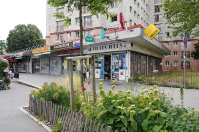 Devanture du commerce Au Fil des Jours (tabac, presse, loto, papeterie) en rez-de-chaussée d’un immeuble d’habitation, avec vitrines remplies de journaux et affiches, enseignes colorées (PMU, tabac, presse) et un trottoir bordé de plantations fleuries au premier plan. - Agrandir l'image, fenêtre modale