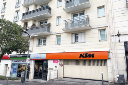 Façade d’un immeuble d’habitation avec balcons, en rez-de-chaussée des commerces dont un magasin de motos et un local signalé par une enseigne orange. Une entrée de parking est visible à droite, donnant sur la rue. - Agrandir l'image, fenêtre modale