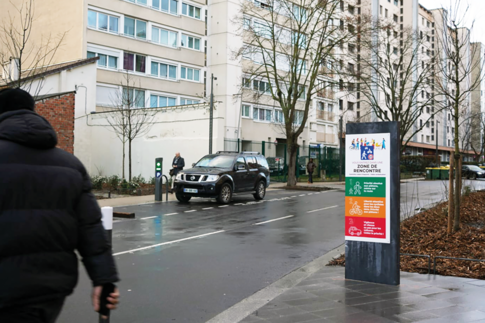 Rue Gabriel Péri, en milieu urbain, avec un panneau signalant l’entrée dans une « zone de rencontre ». Des pictogrammes rappellent le partage de la voirie entre piétons, cyclistes et automobilistes. Un véhicule circule tandis que des passants se déplacent sur les trottoirs, au pied d’immeubles d’habitation.