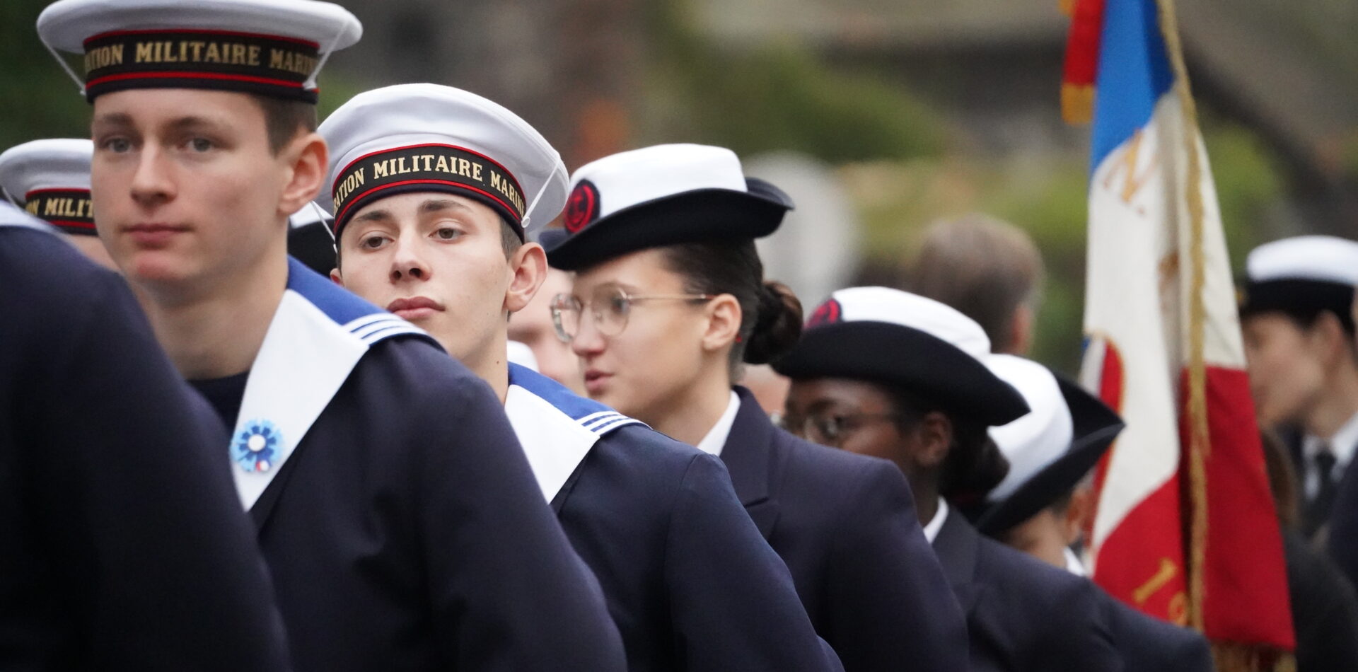 Des élèves en uniforme de la Marine marchent en rang lors d’une cérémonie officielle, avec un drapeau français visible à l’arrière-plan. Au premier plan, plusieurs jeunes portent une vareuse sombre et un bachi blanc marqué « Institution militaire marine ».