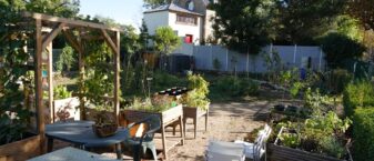 Jardin partagé en milieu urbain, avec des bacs de culture, une pergola en bois, des tables et chaises, entouré de verdure et de quelques habitations en arrière-plan.