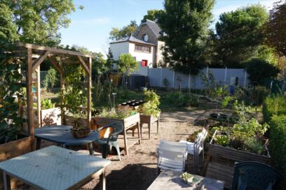 Jardin partagé en milieu urbain, avec des bacs de culture, une pergola en bois, des tables et chaises, entouré de verdure et de quelques habitations en arrière-plan. - Agrandir l'image, fenêtre modale