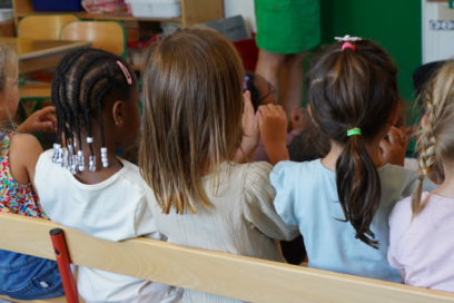 Vue de dos de plusieurs enfants de maternelle assis côte à côte sur un banc dans une salle de classe. Ils regardent attentivement un adulte situé face à eux, hors champ, certains levant la main. L'image illustre un temps d'apprentissage en classe dans une école. - Agrandir l'image, fenêtre modale