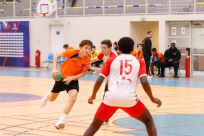 Lors d’un match de handball en salle, un joueur en maillot orange dribble le ballon tout en accélérant vers le but. Deux défenseurs en maillot rouge et blanc tentent de l’arrêter, tandis que des joueurs et entraîneurs observent l’action depuis le banc de touche en arrière-plan. La scène se déroule dans un gymnase éclairé, avec le terrain et ses marquages visibles au premier plan. - Agrandir l'image, fenêtre modale