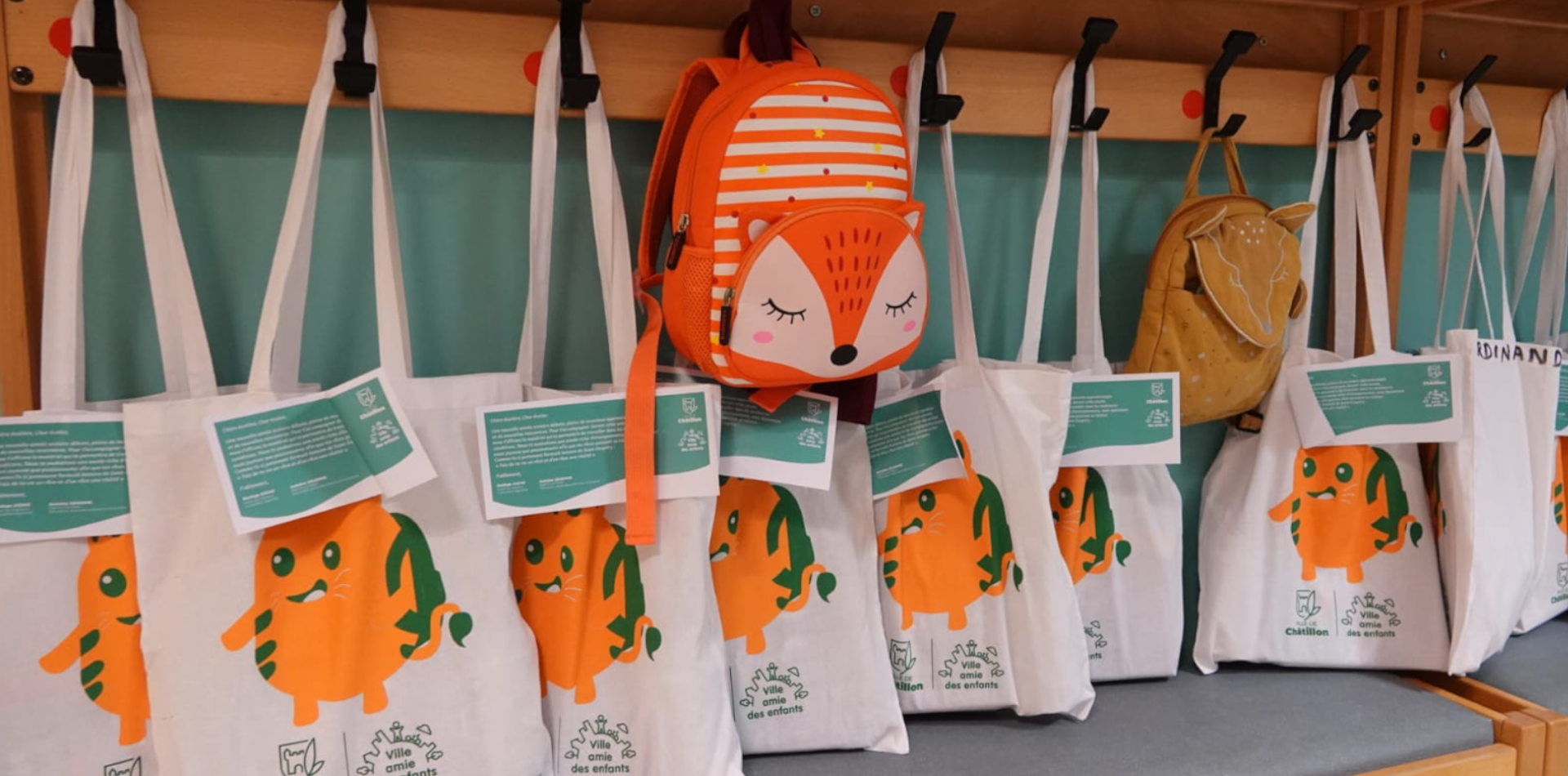 Sacs en tissu blancs aux couleurs de la Ville de Châtillon, accrochés aux porte-manteaux d’une école, aux côtés de sacs à dos d’enfants.