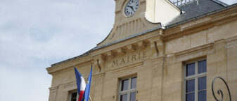 Vue en contre-plongée de la façade de l’Hôtel de Ville de Châtillon, avec son horloge, son inscription « Mairie » et les drapeaux français et européen flottant au-dessus de l’entrée.