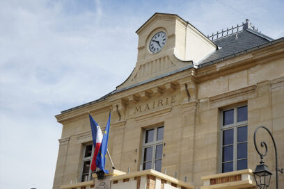 Vue en contre-plongée de la façade de l’Hôtel de Ville de Châtillon, avec son horloge, son inscription « Mairie » et les drapeaux français et européen flottant au-dessus de l’entrée. - Agrandir l'image, fenêtre modale