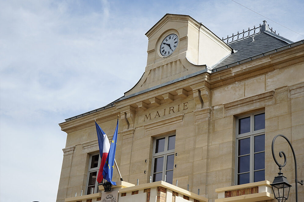 Vue en contre-plongée de la façade de l’Hôtel de Ville de Châtillon, avec son horloge, son inscription « Mairie » et les drapeaux français et européen flottant au-dessus de l’entrée.