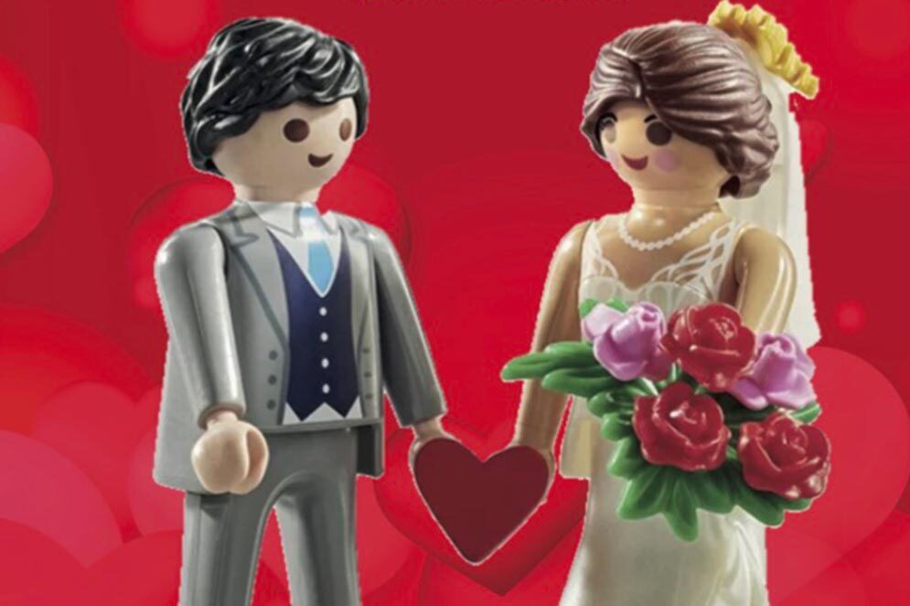 Deux Playmobil, un homme et une femme, se marient. L'épouse est à droite, elle tient un bouquet de fleur, porte une robe blanche et un voile blanc. L'époux, à gauche, porte un costume gris. Ils sont reliés par un coeur qu'ils tiennent par la main.