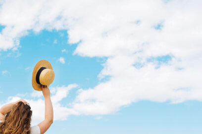 Une femme qui lève son chapeau de paille avec en fond des nuages sur un ciel bleu. - Agrandir l'image, fenêtre modale